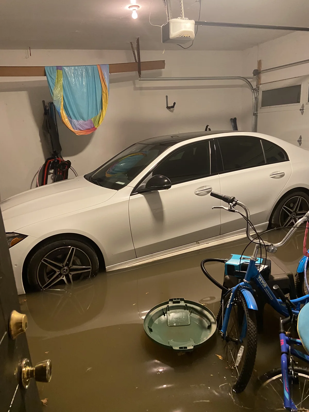 Brown floodwater surrounds a white car and bicycles inside a garage.